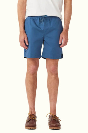 A person wearing a white shirt, RM Williams R.M.W Rugby Shorts in blue cotton with an elasticated waist and a drawstring adjuster, and brown boat shoes stands against a plain background. The shorts feature a casual, relaxed fit.