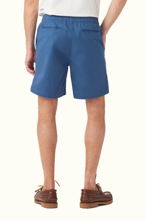 A person wearing the RM Williams R.M.W Rugby Short in blue cotton, featuring an elasticated waist and two back pockets, is standing with their back facing the camera. They are wearing brown shoes, and the background is a plain light gray.