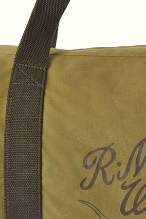 Close-up of the RM Williams RM Ditty Bag in khaki, crafted from waxed cotton canvas with a dark brown handle. The partial view of a black cursive "R" and "M" hints at a printed logo. This stylish accessory from Halley Stevensons is the perfect travel companion.