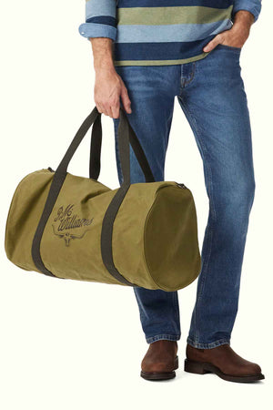 A person in a striped sweater and jeans holds the RM Williams RM Ditty Bag, crafted from green Halley Stevensons waxed cotton canvas. Featuring black straps and a decorative logo, this stylish travel companion is perfectly complemented by their brown shoes.