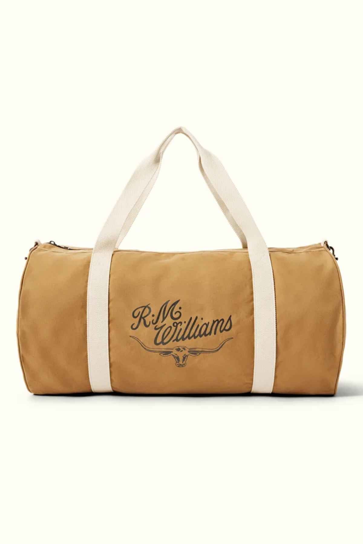 The RM Williams RM Ditty Bag is a tan duffle bag made from waxed cotton canvas, featuring cream handles and an adjustable crossbody strap. The front elegantly showcases "R.M. Williams" along with a decorative graphic, making it your ideal travel companion.