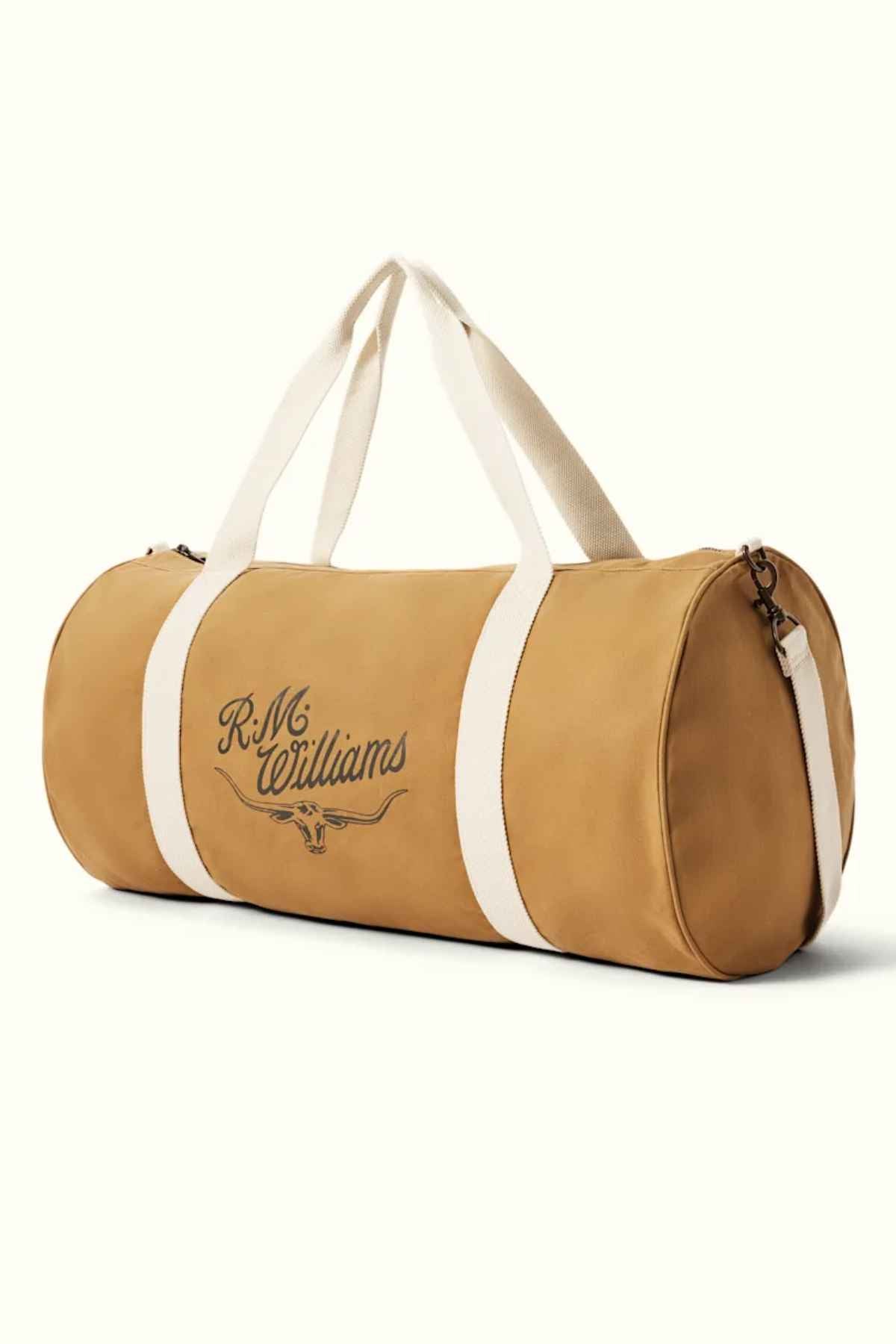 The RM Williams RM Ditty Bag is a tan cylindrical duffle made of waxed cotton canvas, featuring white handles and a zipper. As the perfect travel companion, it displays "R.M. Williams" in a stylish font on the side and includes an adjustable crossbody strap for added convenience.