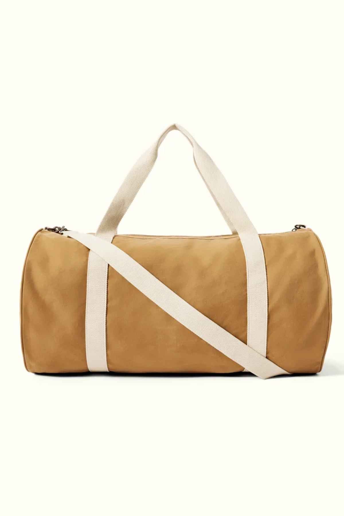 The RM Williams RM Ditty Bag is a tan cylindrical duffel bag crafted from waxed cotton canvas, featuring white straps and an adjustable crossbody strap, making it the perfect travel companion against a plain background.