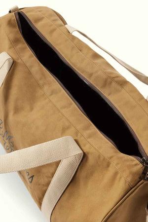 The RM Williams RM Ditty Bag is crafted from brown waxed cotton canvas, featuring a partially open zip that reveals a dark interior. With beige handles and an adjustable crossbody strap, it makes the perfect travel companion. The visible writing on the side enhances its rugged charm.