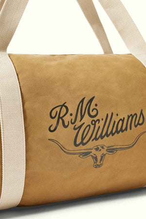 The RM Williams RM Ditty Bag is displayed, a tan duffel with white straps that makes an ideal travel companion. It showcases the R.M. Williams logo in black on the side, featuring a cattle head with horns. Crafted from leather or leather-like material, it includes an adjustable crossbody strap for added convenience while traveling.