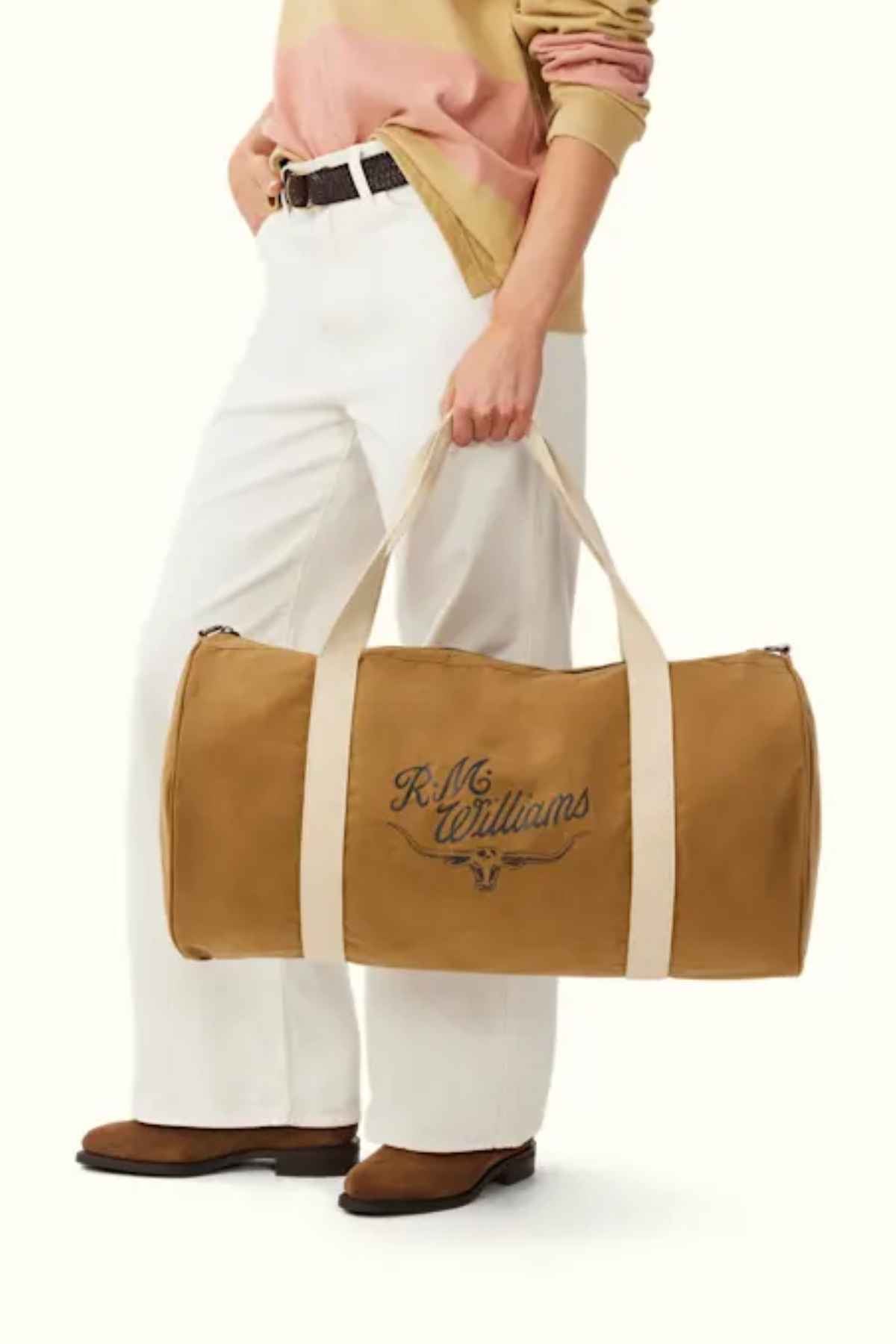 A person dressed in a light pink and brown sweater matched with white pants stylishly holds the RM Williams RM Ditty Bag, made from tan waxed cotton canvas. The bag features beige straps, an adjustable cross-body strap, and a recognizable logo, perfectly complemented by brown shoes.