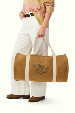 A person dressed in a light pink and brown sweater matched with white pants stylishly holds the RM Williams RM Ditty Bag, made from tan waxed cotton canvas. The bag features beige straps, an adjustable cross-body strap, and a recognizable logo, perfectly complemented by brown shoes.