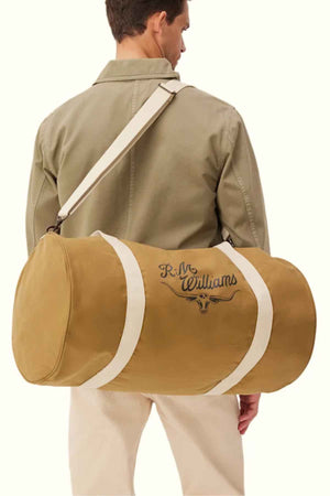 A person dressed in a khaki shirt and cream pants carries the RM Williams RM Ditty Bag, a large duffle bag featuring white straps and an adjustable cross-body strap, ideal as a travel companion. The tan bag is crafted from waxed cotton canvas and proudly displays the "RM Williams" logo alongside a distinctive bull design.