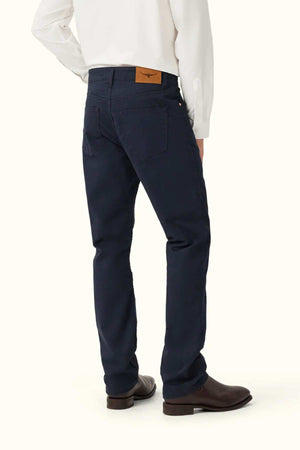 A person wearing RM Williams Ramco 5 Pocket Jeans in dark blue Australian cotton, brown boots, and a white long-sleeve shirt stands facing away, revealing the jeans’ brown leather waistband patch.