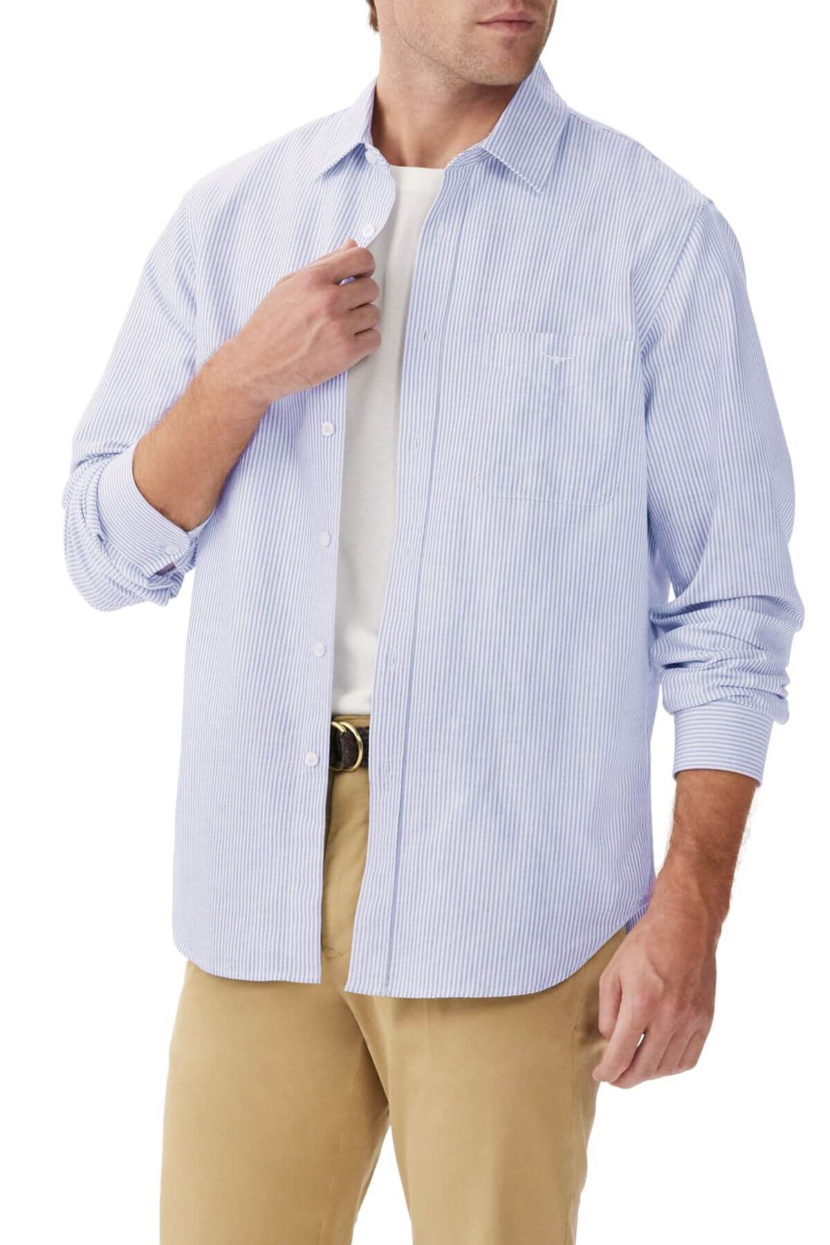 Wearing the RM Williams Regular Shirt in light blue and white stripes over a white t-shirt, paired with beige pants. Made from organic cotton, the long-sleeve shirt, rolled to the elbows, is adjusted at the collar by hand.