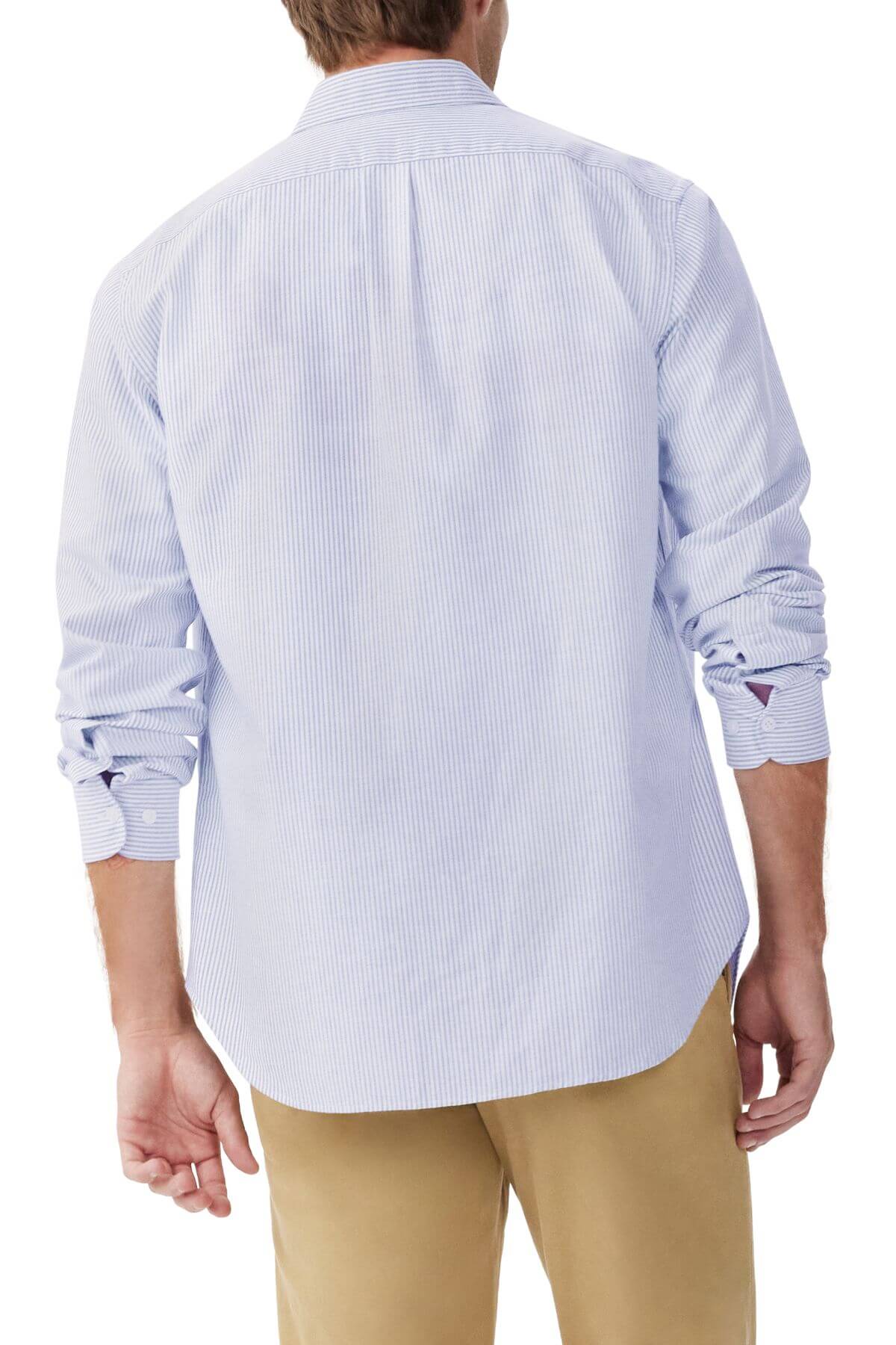 A person is wearing the RM Williams Regular Shirt, a slim fit light blue pinstripe button-up crafted from organic cotton, paired with beige pants. The shirt's sleeves are rolled up to the elbows, showcasing casual elegance against a plain white backdrop.