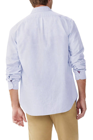 A person is wearing the RM Williams Regular Shirt, a slim fit light blue pinstripe button-up crafted from organic cotton, paired with beige pants. The shirt's sleeves are rolled up to the elbows, showcasing casual elegance against a plain white backdrop.