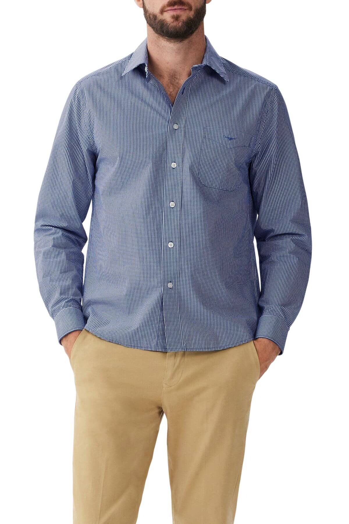 A person is wearing an RM Williams Classic Shirt in a blue and white micro gingham check pattern with long sleeves, paired with beige trousers. Their hands are in their pockets, and the image is taken from the knees up. The background is plain and unobtrusive.