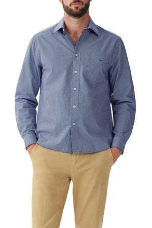 A person is wearing an RM Williams Classic Shirt in a blue and white micro gingham check pattern with long sleeves, paired with beige trousers. Their hands are in their pockets, and the image is taken from the knees up. The background is plain and unobtrusive.