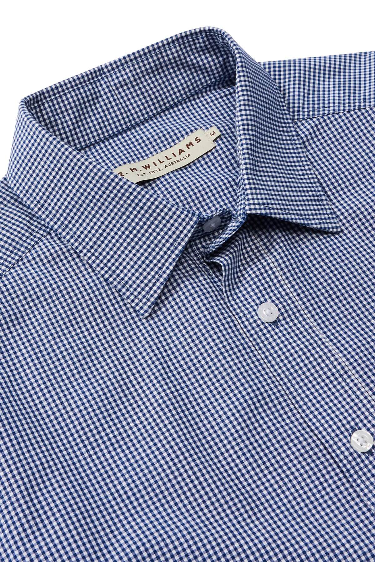 Close-up of a neatly folded RM Williams Classic Shirt, featuring a blue and white micro gingham check pattern. This regular fit, button-up shirt has a pointed collar, is made from organic cotton, and includes white buttons with a visible label at the neck displaying text and details.