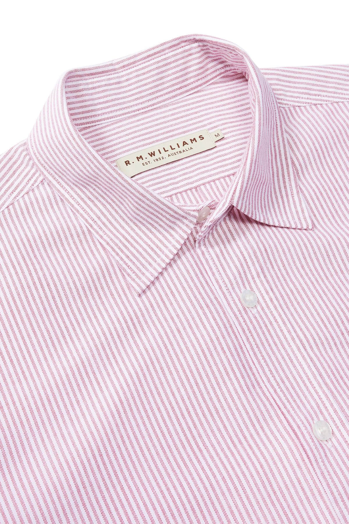 A close-up of the RM Williams Regular Shirt, featuring light pink and white stripes, a collared button-down style, and a medium "R.M. Williams" tag. Made from organic cotton for comfort in a slim regular fit.