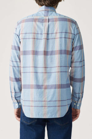 A person is seen from behind wearing RM Williams' regular fit shirt in light blue plaid, featuring light orange and gray stripes, paired with dark blue jeans. The shirt is made of organic cotton. They stand against a plain light gray background.