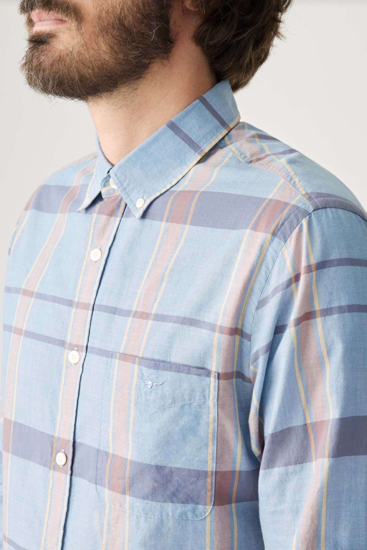 A person is wearing the RM Williams Regular Shirt, featuring a blue plaid design with stripes of blue, orange, and cream. It has a button-down collar and an embroidered logo on the chest pocket. Their beard and part of their face are visible.