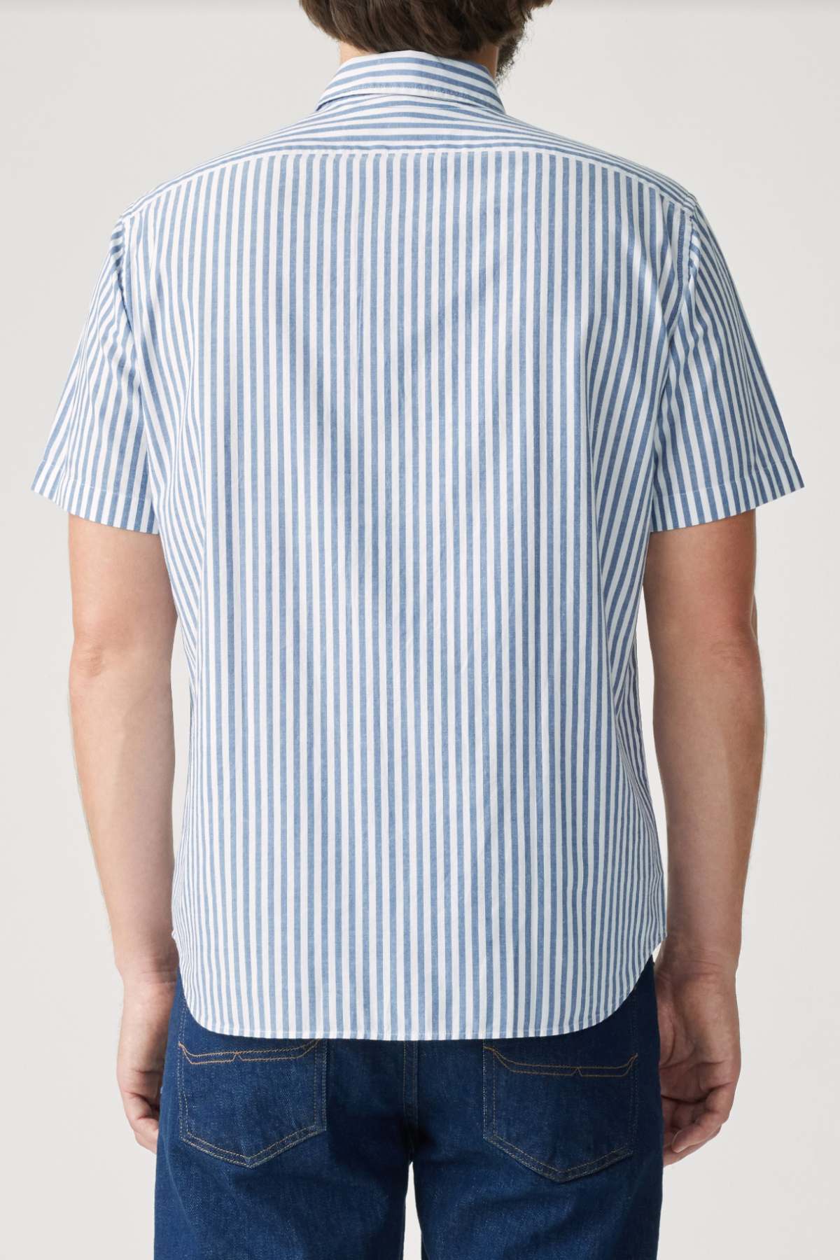A person is seen from the back wearing the RM Williams Regular Short Sleeve Shirt with blue and white vertical stripes, a button-down collar, and dark blue jeans.