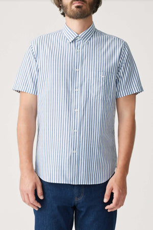 A man wearing an RM Williams Regular Short Sleeve Shirt with vertical blue and white stripes and a button-down collar, paired with dark blue jeans, stands against a plain light background.