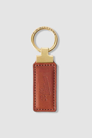 The RM Williams Ridgewood Key Fob has a rectangular brown leather strap, a gold-colored keyring, and is embossed with cursive text. This elegant and timeless design adds sophistication to key organization.