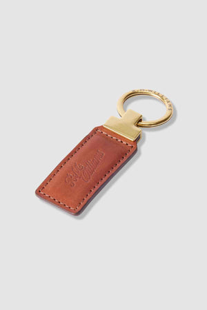 R.M. Williams Ridgewood Key Fob: This elegant gold keyring features a rectangular brown leather tag with the R.M. Williams logo embossed on it. Detailed edge stitching enhances its classic look, making it stand out beautifully against a plain white background.