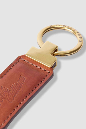 A close-up of the RM Williams Ridgewood Key Fob reveals embossed text on rich brown leather, a gold-tone metal ring with the R.M. Williams logo, and a sleek gold connector, exuding timeless sophistication.