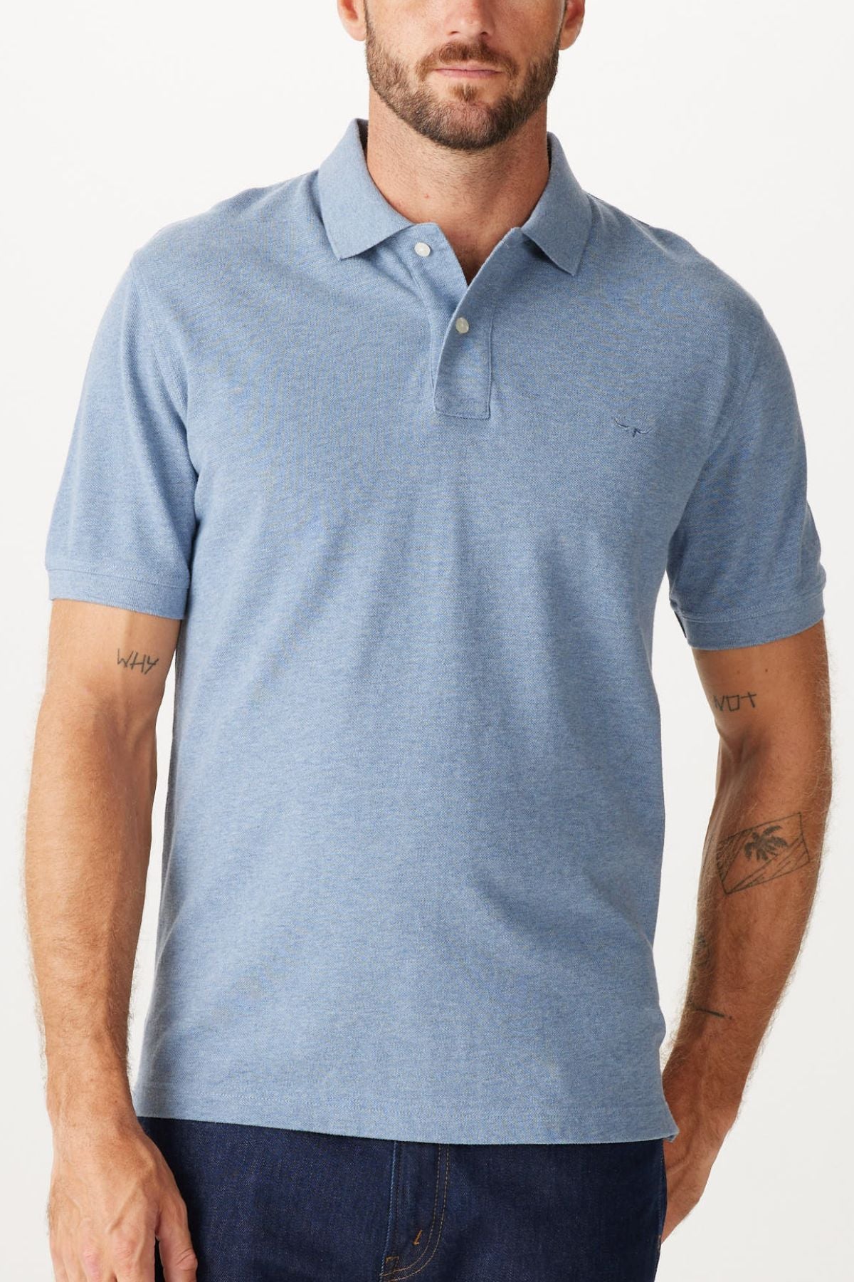 A man wearing the RM Williams Rod Polo, a light blue cotton pique shirt with collar, two buttons, and subtle Longhorn chest embroidery stands against a plain white background. He pairs it with dark jeans, his tattooed forearms on display.