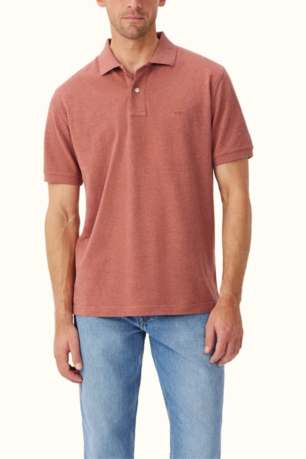 A man wearing the RM Williams Rod Polo in a rust color, designed with a classic fit from organic cotton, stands against a plain background. The light blue jeans accentuate the polo's subtle embroidered logo on the chest.