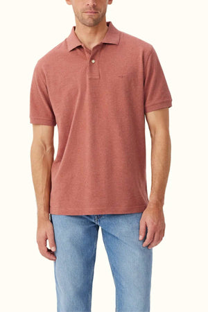 A man wearing the RM Williams Rod Polo in a rust color, designed with a classic fit from organic cotton, stands against a plain background. The light blue jeans accentuate the polo's subtle embroidered logo on the chest.