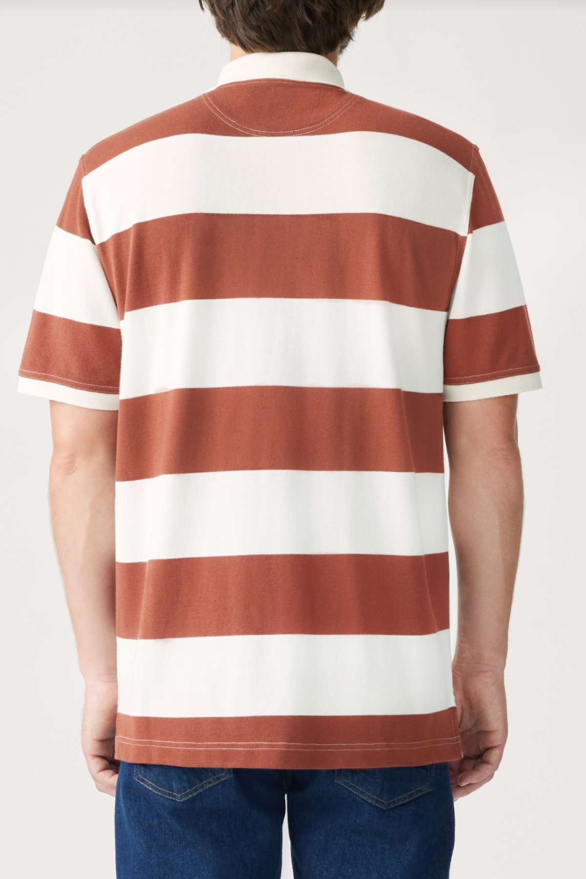 Rear view of a person wearing the RM Williams Rod Polo, a short-sleeved, red and white striped shirt made from breathable cotton, paired with blue jeans, standing against a plain background.