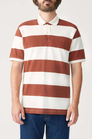 A person in the RM Williams Rod Polo, a regular fit polo shirt with short sleeves and featuring broad horizontal stripes in brown and white. This breathable cotton shirt is adorned with an embroidered longhorn on the chest and completed with a classic collar, paired perfectly with blue jeans, as they stand against a plain background.
