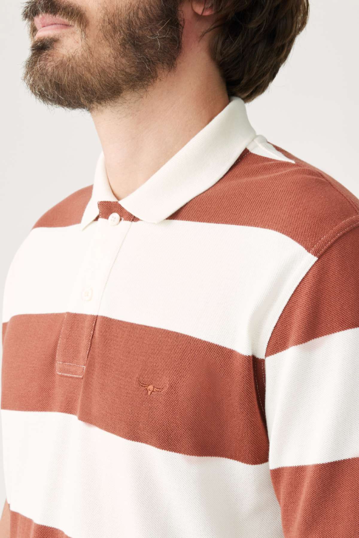 A man in the RM Williams Rod Polo, featuring a breathable fabric and a brown and white striped pattern, showcases an embroidered longhorn logo on the chest, emphasizing his beard and the regular fit of the shirt.