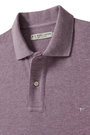 A close-up of the mauve RM Williams Rod Polo showcases its folded collar, two-button placket, small longhorn chest embroidery, and visible brand label inside the collar. The shirt is crafted from breathable cotton pique fabric.