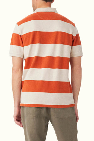 The image displays a person dressed in the RM Williams Rod Polo, a short-sleeved, orange and beige striped shirt crafted from organic cotton, seen from the back. They pair it with olive green classic-fit pants against an off-white background.