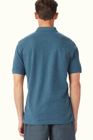 A person wearing a RM Williams Rod Polo in a classic fit blue style is shown from the back. They have short hair and are paired with gray shorts made from organic cotton, set against a plain off-white background.
