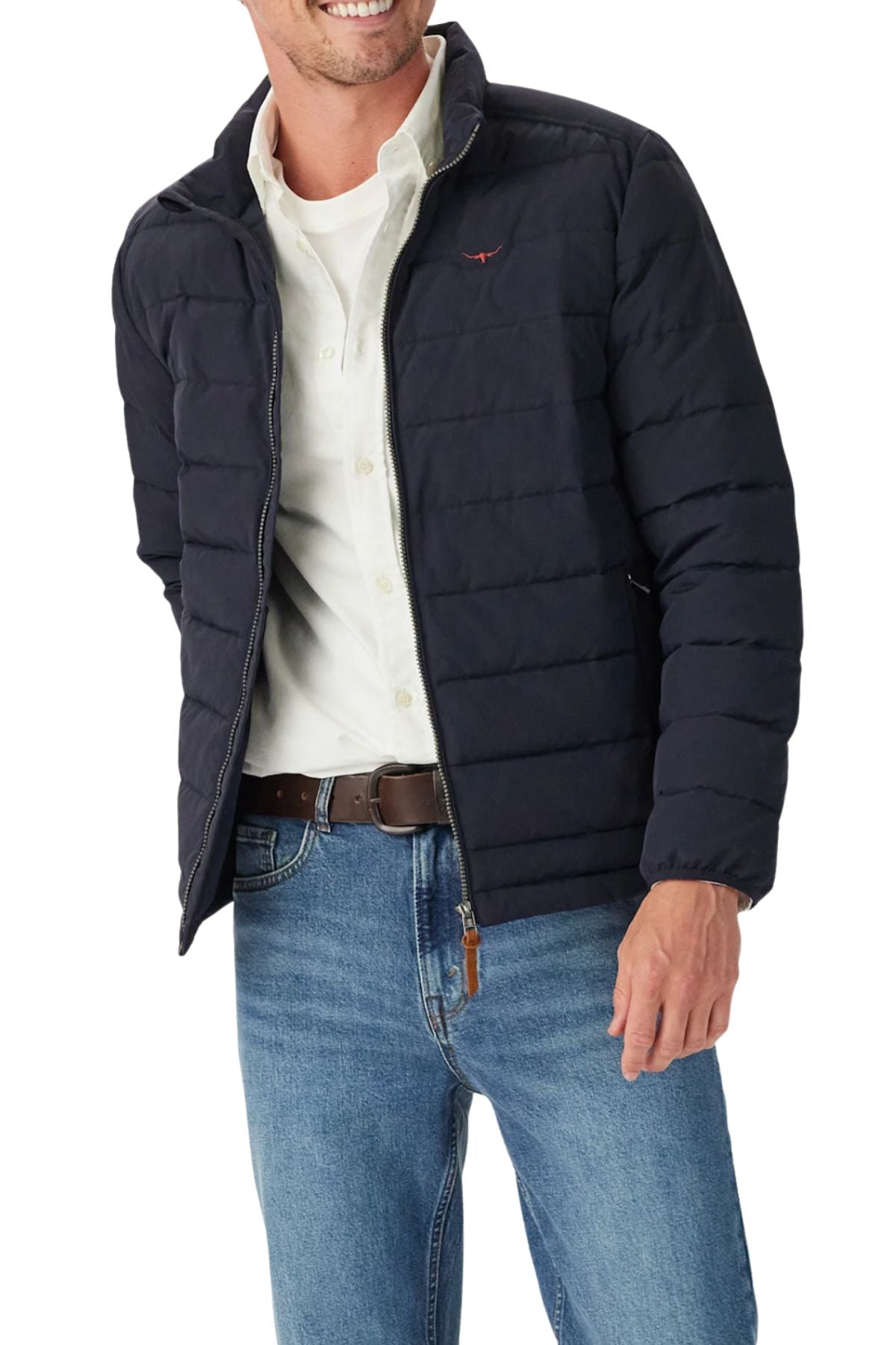 A man wears the RM Williams Rydal Padded Jacket over a white button-down shirt, blue jeans, and a brown belt. He’s smiling with his right hand in his pocket.
