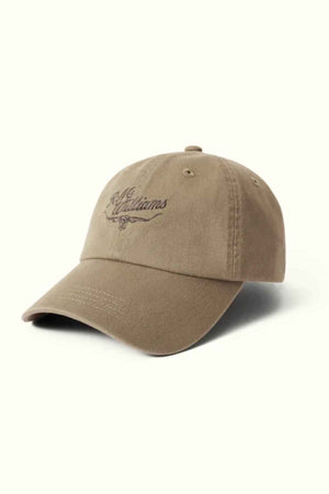 The RM Williams Script Longhorn Cap is a brown baseball cap embroidered with "The Mishum" in dark brown thread, showcasing a heritage script logo. Made from washed cotton twill, it features a curved brim and visible stitching on a light background.