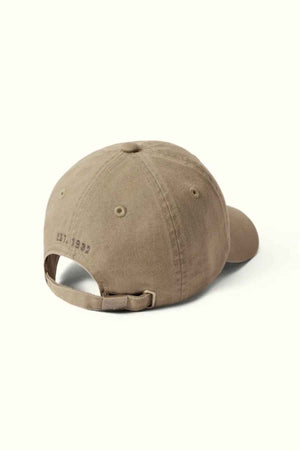 Viewed from the back, the RM Williams Script Longhorn Cap is a brown baseball cap made from washed cotton twill. It features an adjustable strap and subtly embroidered text reading "EST. 1992." Its simple design and neutral color lend it a casual look.