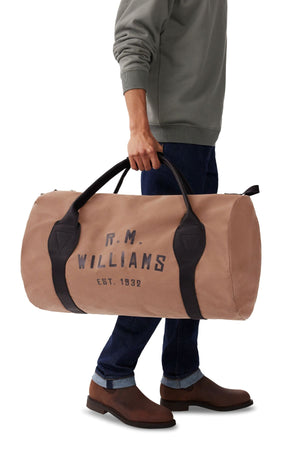 A person in a grey sweatshirt, blue jeans, and brown boots holds the RM Williams Sorrento Ute Bag with black straps labeled "R.M. Williams Est. 1932." Only the lower half of their body is visible.