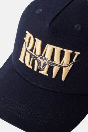 The RM Williams Steers Head Trucker Cap is a navy blue hat featuring gold embroidered "RMW" lettering and a central longhorn skull design, equipped with an adjustable snapback for an ideal fit.