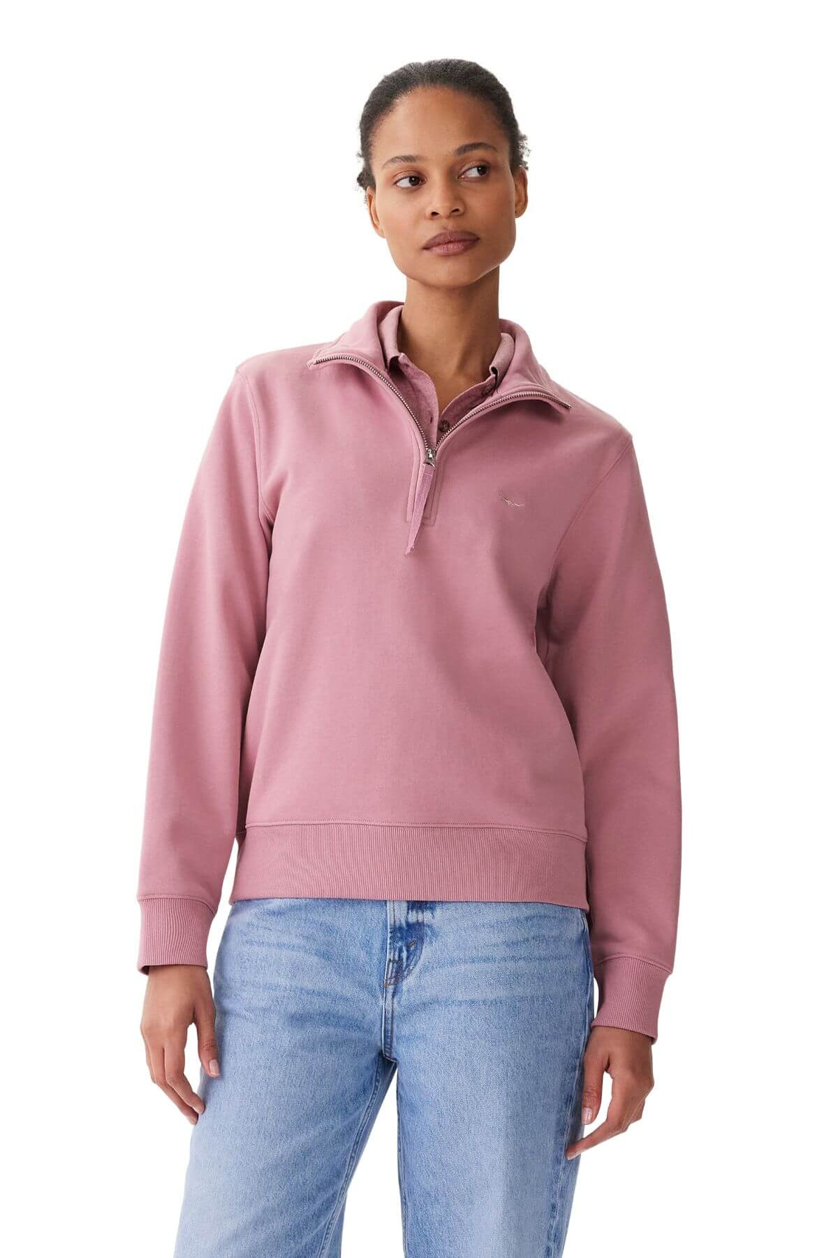 A person in an RM Williams Trickett Zip Neck Sweatshirt with a longhorn embroidery and light blue jeans stands against a plain white background, short hair framing their face as they gaze thoughtfully to the side.