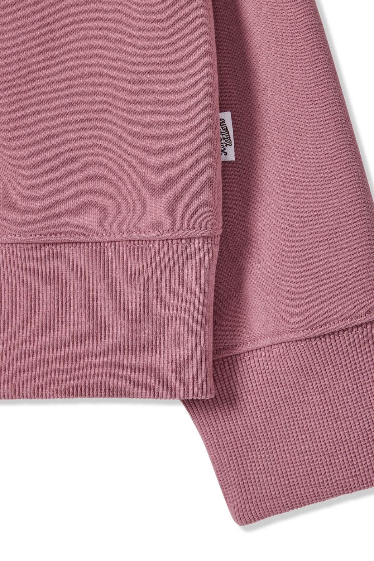 Close-up of the RM Williams Trickett Zip Neck Sweatshirt in pink, made from loop back cotton terry with ribbed cuffs and hem. A small white fabric tag with text is attached to the side, and the material appears soft and textured.