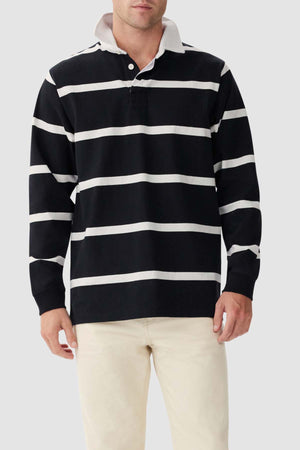 A man is standing against a plain background wearing the RM Williams Tweedale Rugby jersey, which features black and white horizontal stripes and a white collar. He is also in light-colored pants. His face is partially visible, and his hands are by his sides.
