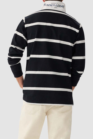 A person wearing the RM Williams Tweedale Rugby, a black heavyweight knit shirt with white horizontal stripes and a white collar, paired with light-colored pants. The back of the shirt features the text "Australia R. M. Williams" in white and blue. The person is standing against a gray background.