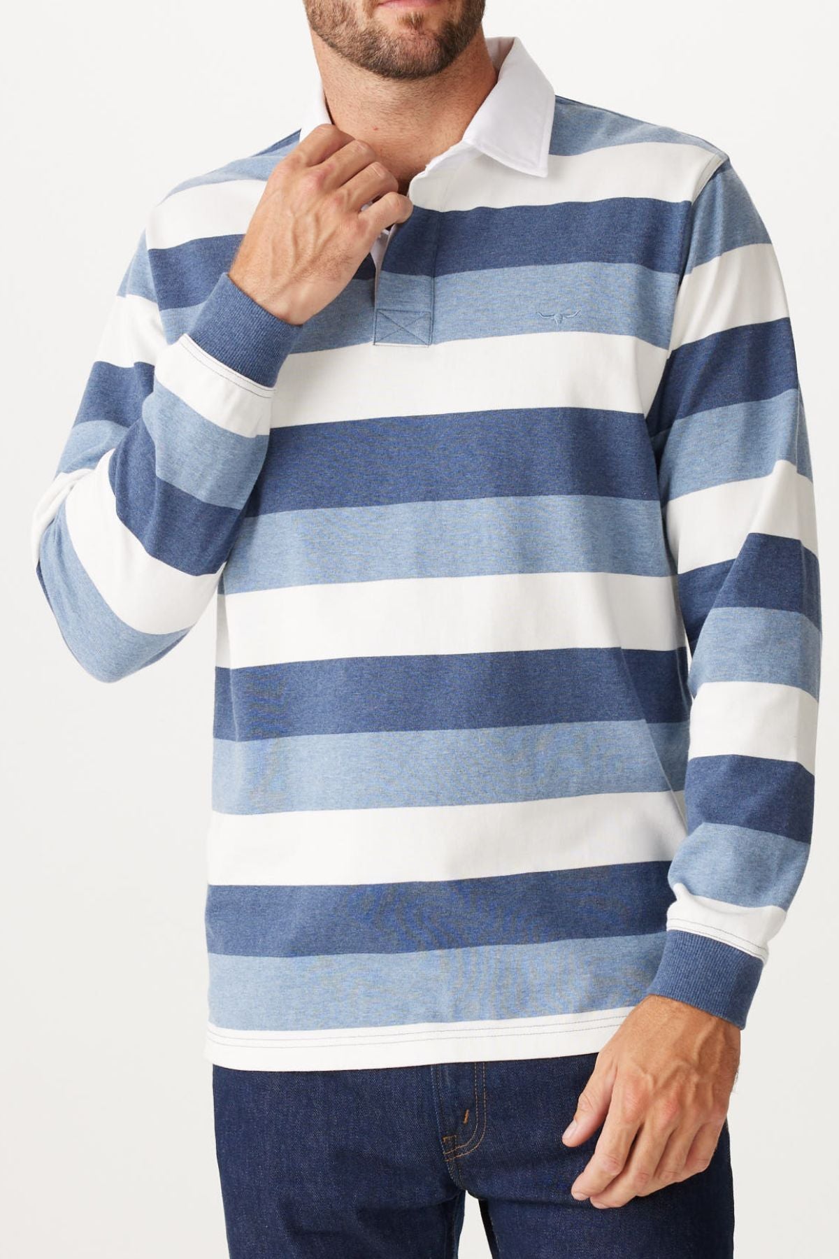 A man is wearing the RM Williams Tweedale Rugby shirt, made from heavyweight jersey with bold blue and white stripes. He's adjusting the collar with one hand and pairing it with dark jeans for a timeless look.