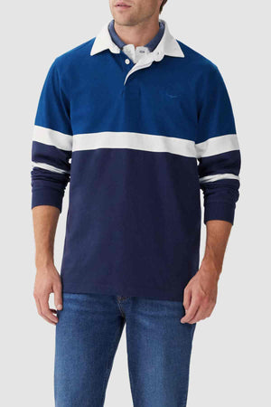A person wearing the RM Williams Tweedale Rugby, a heavyweight blue rugby shirt featuring longhorn embroidery on the chest and a white collar. The person is also wearing blue jeans, set against a plain and light-colored background.