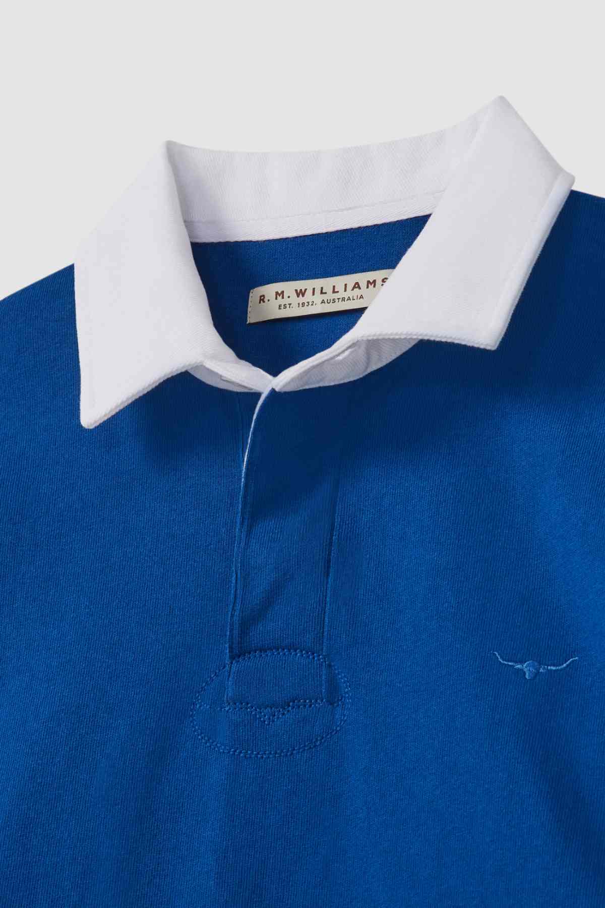 Close-up of the RM Williams Tweedale Rugby, a blue heavyweight jersey rugby shirt with a white collar. It features a small, stitched longhorn embroidery on the left side of the chest and a label inside the collar that reads "R.M. Williams Est. 1932 Australia.