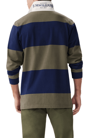 A person shown from behind wears the RM Williams Tweedale Rugby—a heavyweight jersey rugby shirt with bold navy and olive green stripes, a white collar printed with "Australia R.M. Williams Est. 1932"—paired with olive green pants.