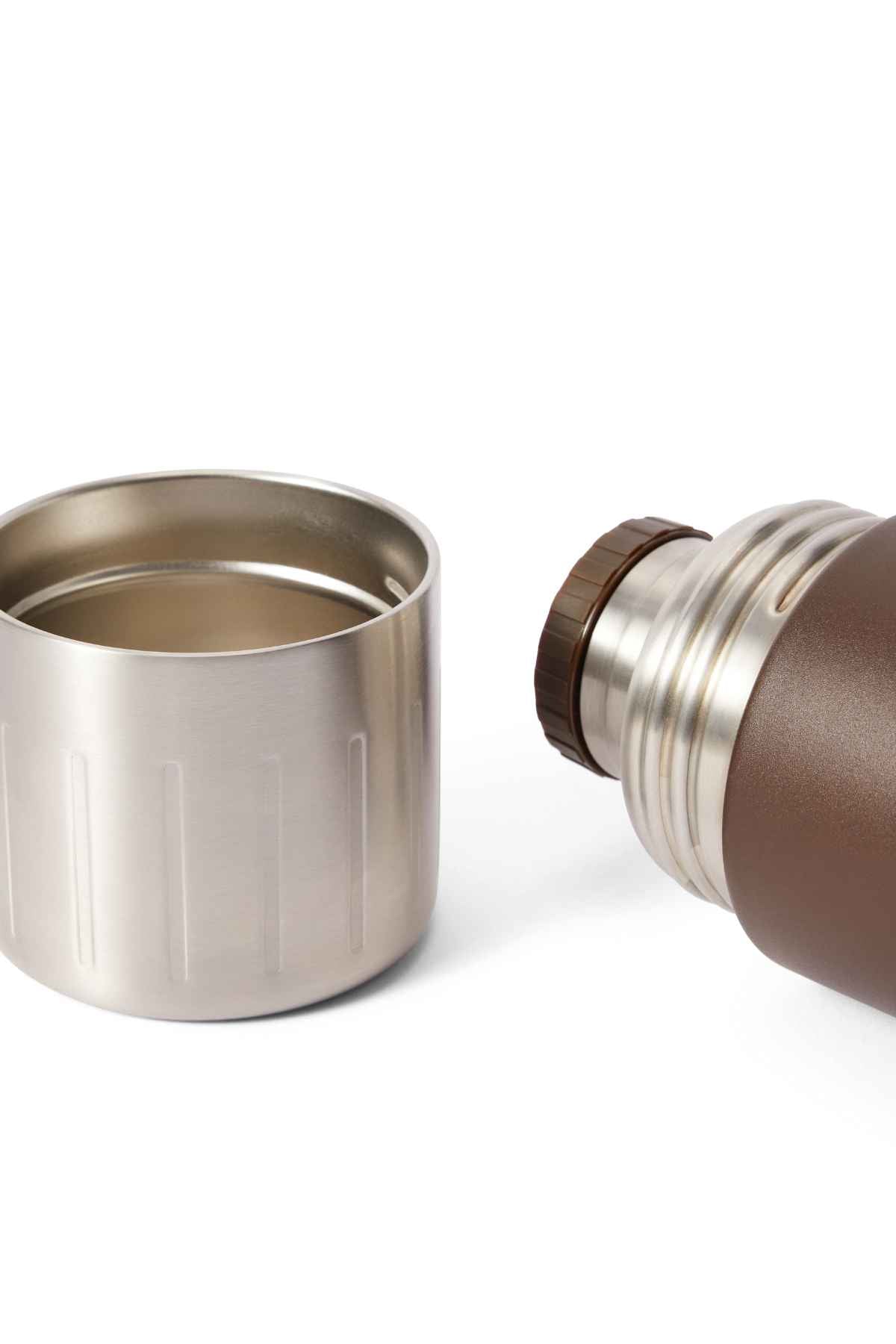 The RM Williams Vacuum Flask, made of stainless steel, lies on its side without its cap. The brushed metal cap, doubling as a cup, is to the left. This brown thermos has double wall vacuum insulation and a metallic section near the opening for durability.